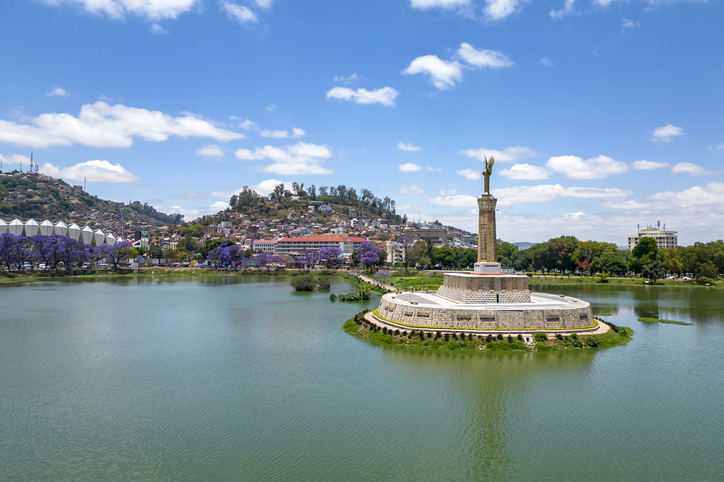 Inmitten eines kreisrunden Sees steht eine goldene Engelsstatue auf einem hohen Steinsockel, umgeben von blühenden lila Jacaranda-Bäumen.