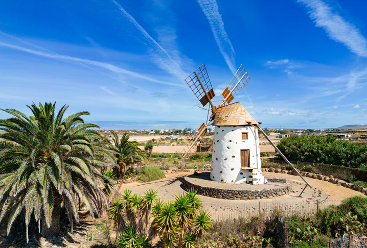Eine weiße, konische Windmühle mit hölzernen Flügeln steht auf einem runden Steinsockel vor einem weiten Feld und blauem Himmel.