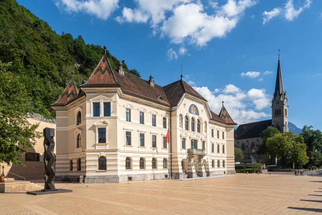 Regierungsgebäude mit Liechtenstein-Flagge und Kirchturm vor Bergen.