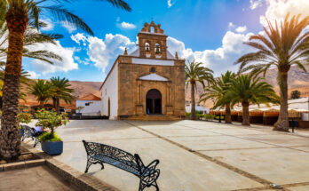Eine weiß getünchte Kapelle mit Natursteinfassade steht an einem sonnigen Platz, gesäumt von Palmen und schmiedeeisernen Bänken.
