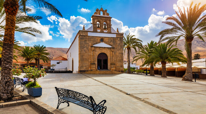 Eine weiß getünchte Kapelle mit Natursteinfassade steht an einem sonnigen Platz, gesäumt von Palmen und schmiedeeisernen Bänken.
