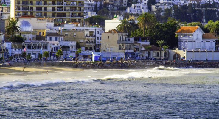 Ein langer Sandstrand mit brechender Brandung liegt direkt vor einer Kulisse aus modernen Hochhäusern und historischen Villen.