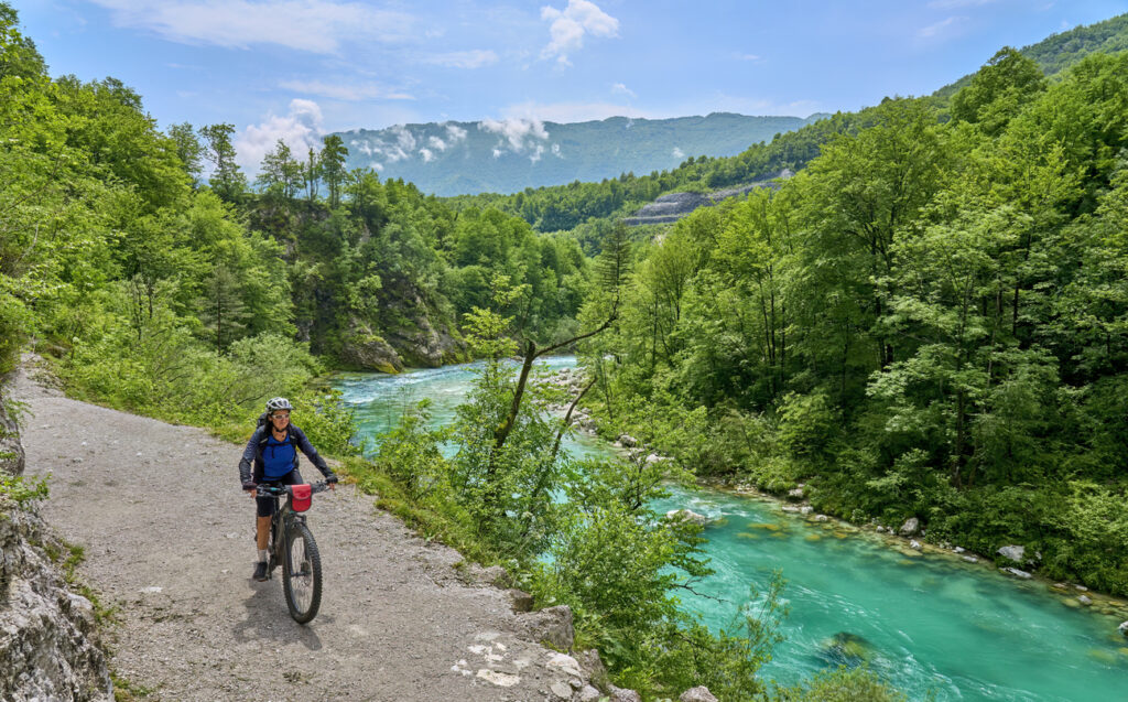 Eine Radfahrerin fährt auf einem Schotterweg entlang eines klaren, türkisgrünen Flusses durch dichten Wald.