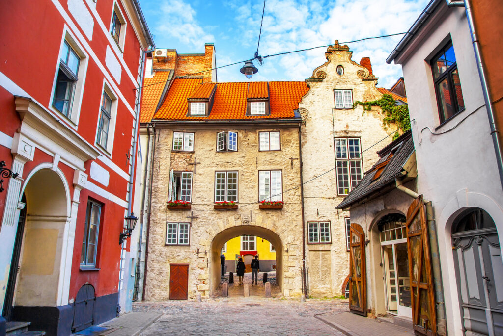 Ein gepflasterter Weg führt durch einen Torbogen in einem historischen sandsteinfarbenen Gebäude.