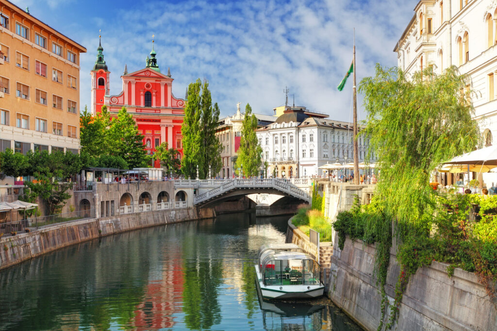 Ein Ausflugsboot liegt am Ufer des Flusses in Ljubljana unterhalb der berühmten Drei Brücken und der markanten rosa Kirche.