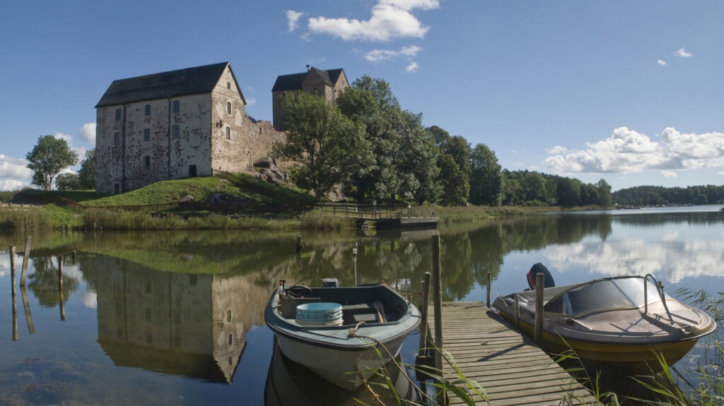 Zwei kleine Boote liegen an einem Holzsteg vor einem ruhigen Gewässer, während sich ein altes Steingebäude im Wasser spiegelt.
