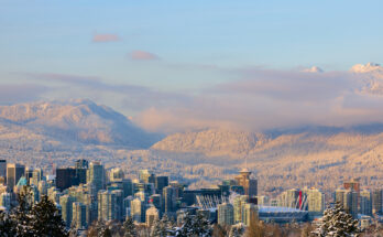 Ein weiter Blick über die Skyline von Vancouver auf die schneebedeckten North Shore Mountains unter einem zartrosa Abendhimmel.