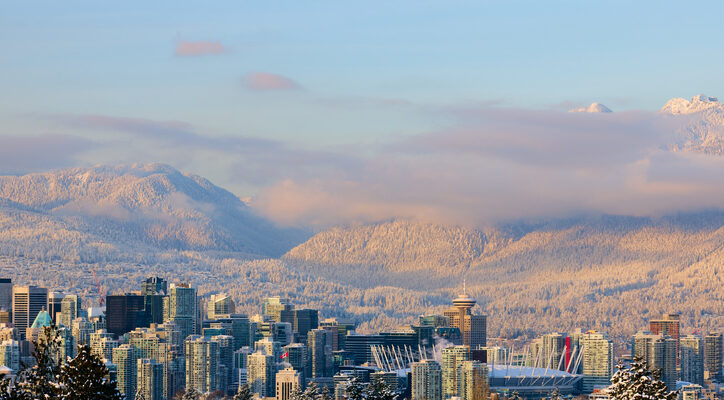 Ein weiter Blick über die Skyline von Vancouver auf die schneebedeckten North Shore Mountains unter einem zartrosa Abendhimmel.