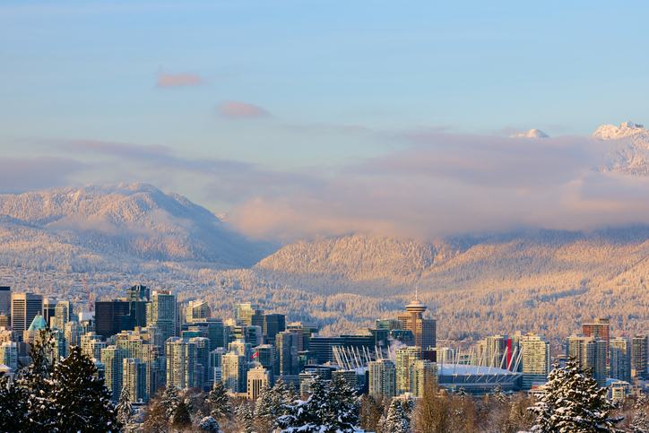 Ein weiter Blick über die Skyline von Vancouver auf die schneebedeckten North Shore Mountains unter einem zartrosa Abendhimmel.