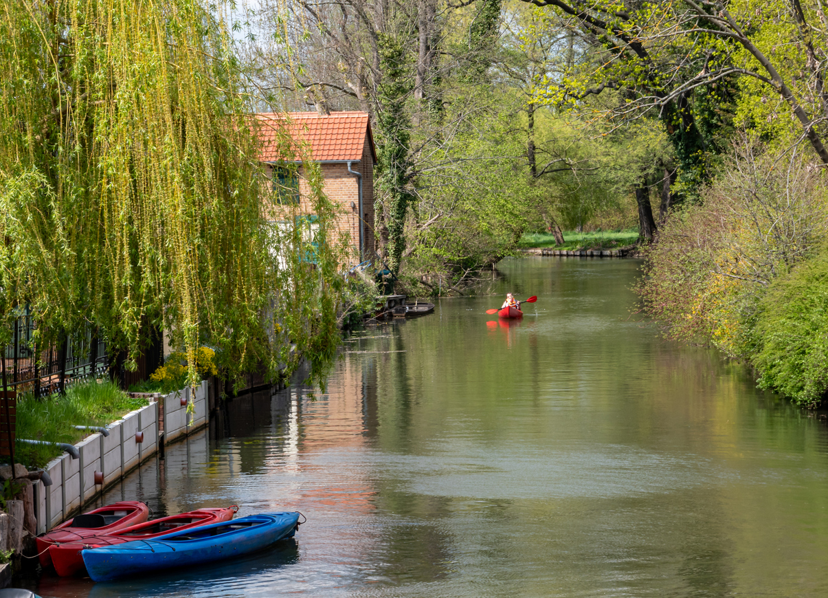 Spreewaldkanal mit Bäumen, Häusern am Ufer und einem roten Kajak im Hintergrund.