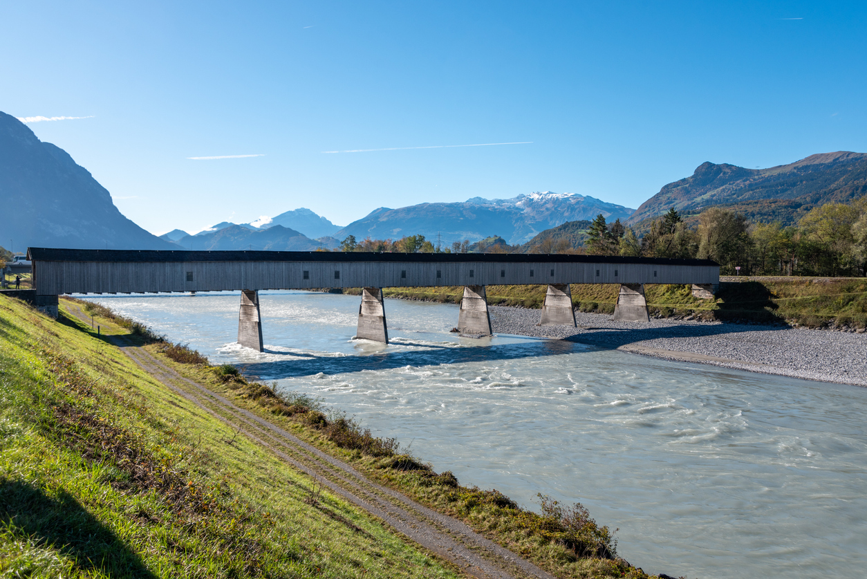 Lange Holzbrücke über einem breiten Fluss mit Bergen im Hintergrund.
