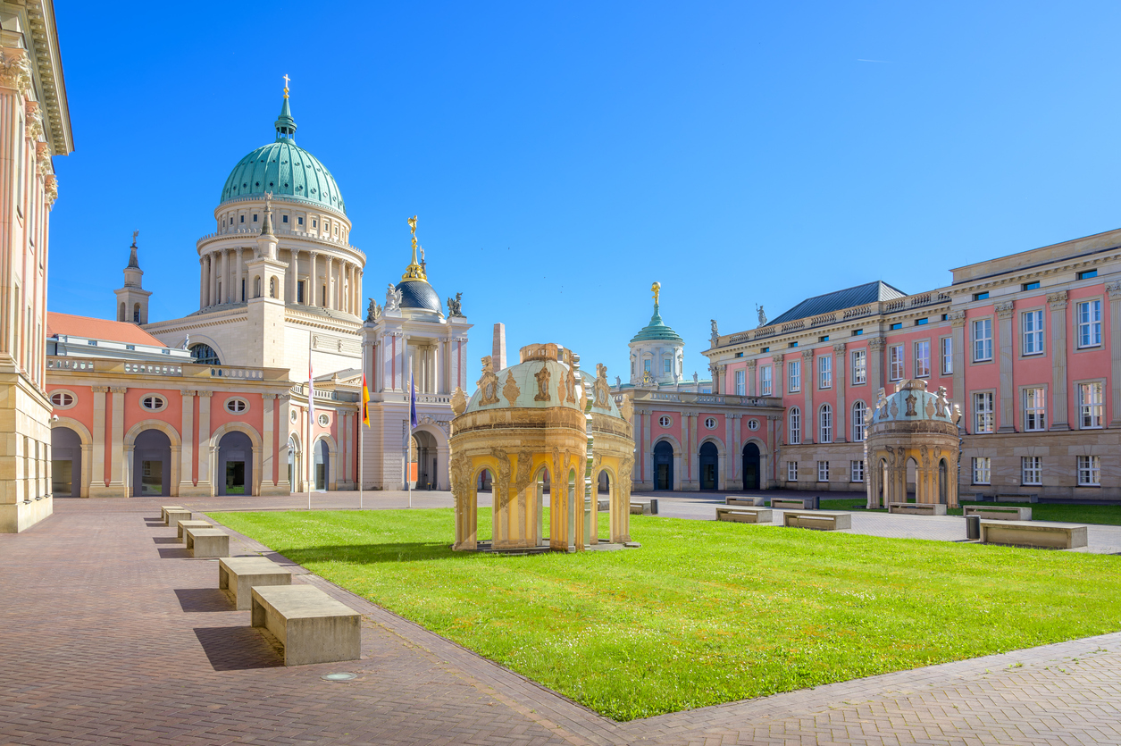 Die imposante Barockfassade des Neuen Palais mit seiner großen Kuppel und den markanten, geschwungenen Treppenaufgängen vor einem blauen Himmel.