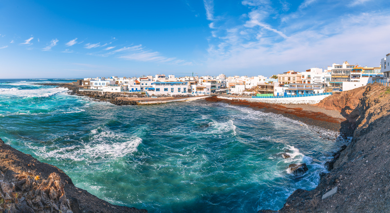 Eine Bucht mit tiefblauem, unruhigem Wasser liegt vor einer Ansammlung weißer Häuser mit blauen Akzenten unter strahlendem Himmel.
