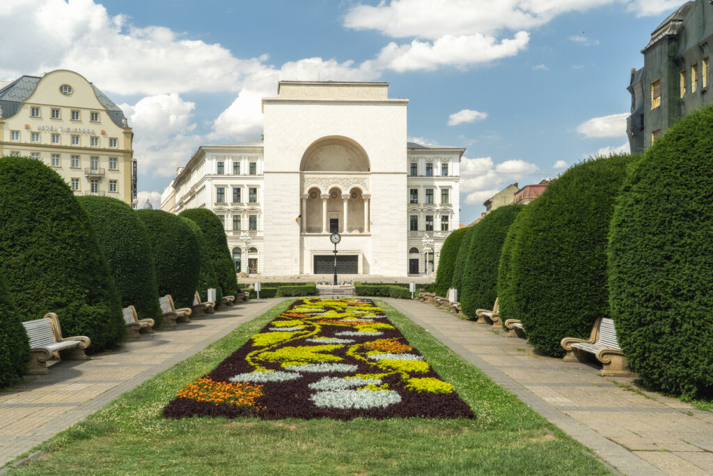 Ein imposantes, weißes Gebäude mit einer großen zentralen Bogenöffnung und Säulen steht am Ende eines Parks mit blühenden Blumenbeeten.