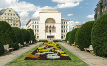 Ein imposantes, weißes Gebäude mit einer großen zentralen Bogenöffnung und Säulen steht am Ende eines Parks mit blühenden Blumenbeeten.