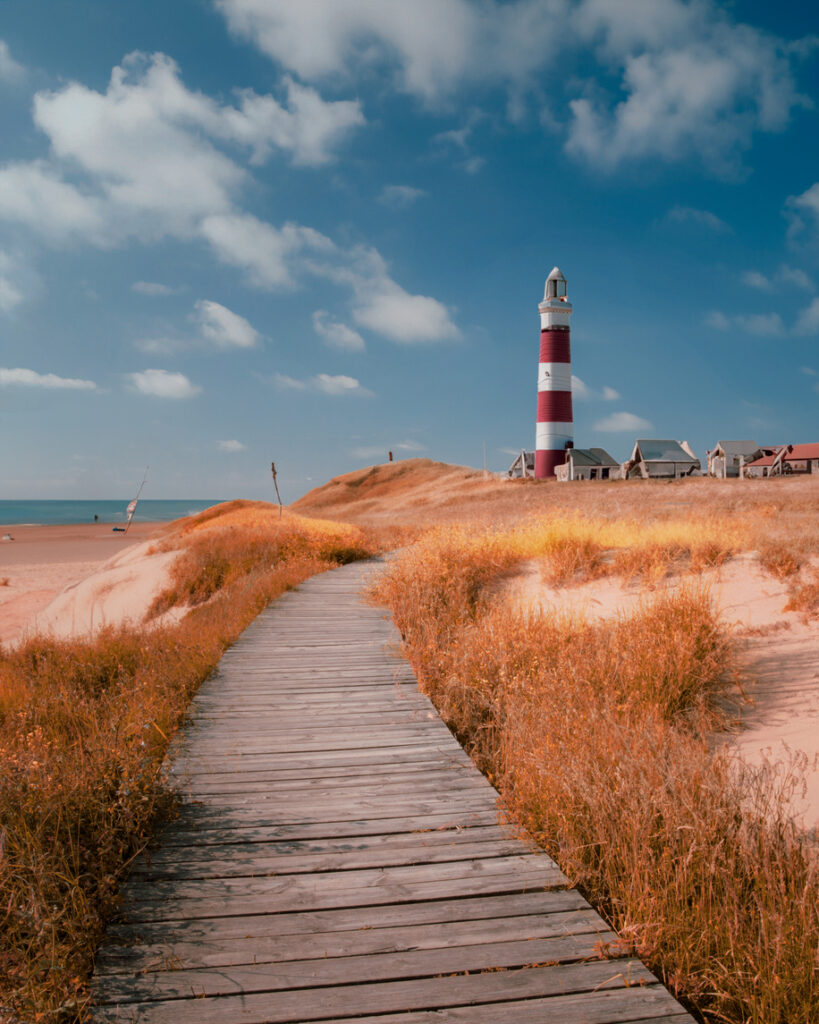 Ein rot-weiß gestreifter Leuchtturm steht am Ende eines hölzernen Bohlenweges, der durch eine Dünenlandschaft führt.