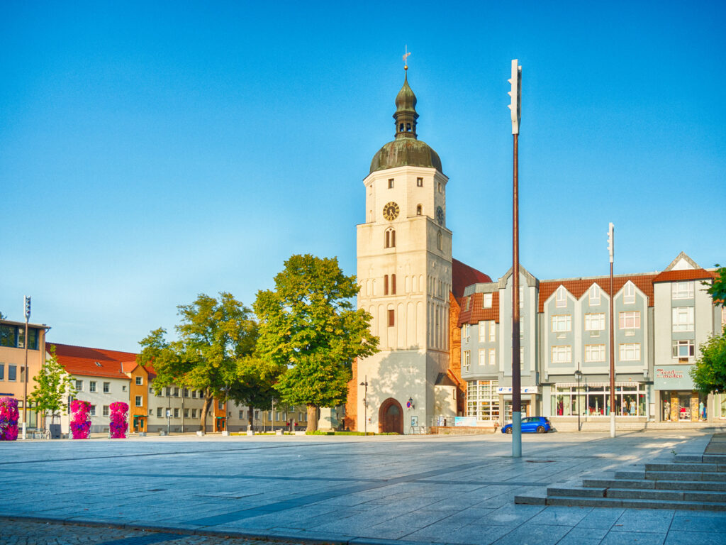 Großer Platz mit Kirchturm und Häuserzeile bei blauem Himmel.