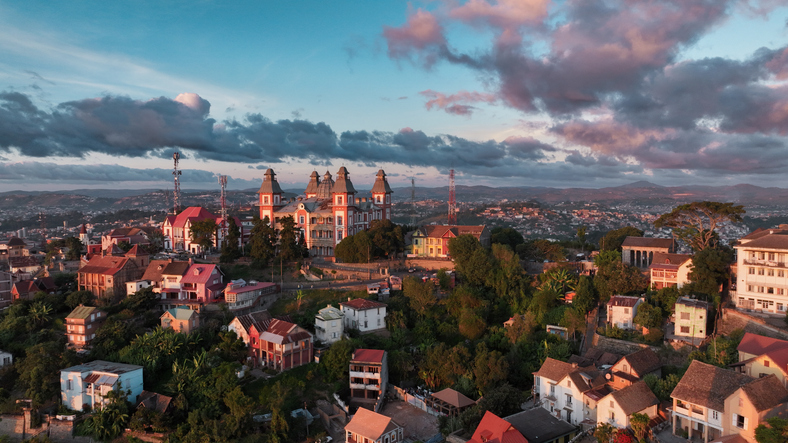 Ein herrschaftliches, rot-weißes Gebäude mit Türmen steht auf einem bewaldeten Hügel über zahlreichen Wohnhäusern unter einem dramatischen Wolkenhimmel.