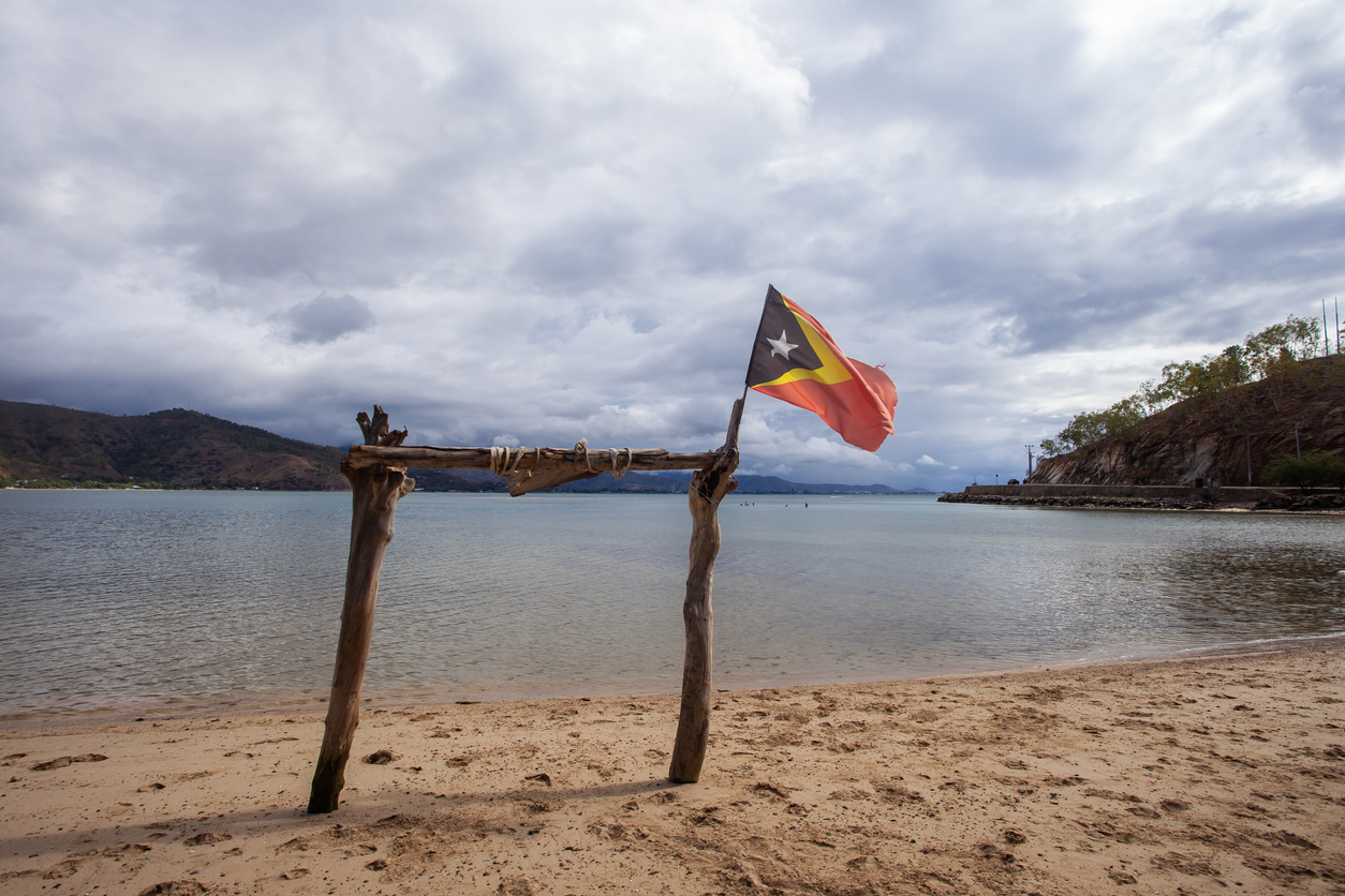 Am Strand in Osttimor steht ein Holzgestell mit wehender Flagge vor einer stillen Bucht und bewölktem Himmel.