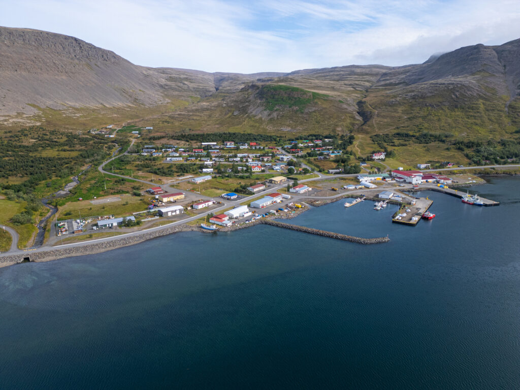Luftaufnahme eines kleinen Ortes mit Hafen, Booten und Bergen an einer Küste in den Westfjorden auf Island.