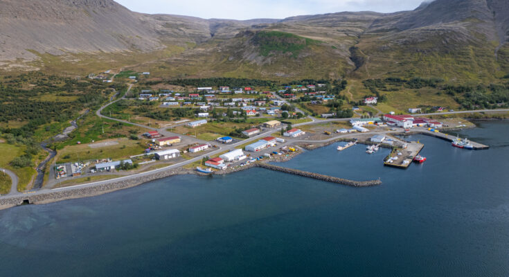 Luftaufnahme eines kleinen Ortes mit Hafen, Booten und Bergen an einer Küste in den Westfjorden auf Island.