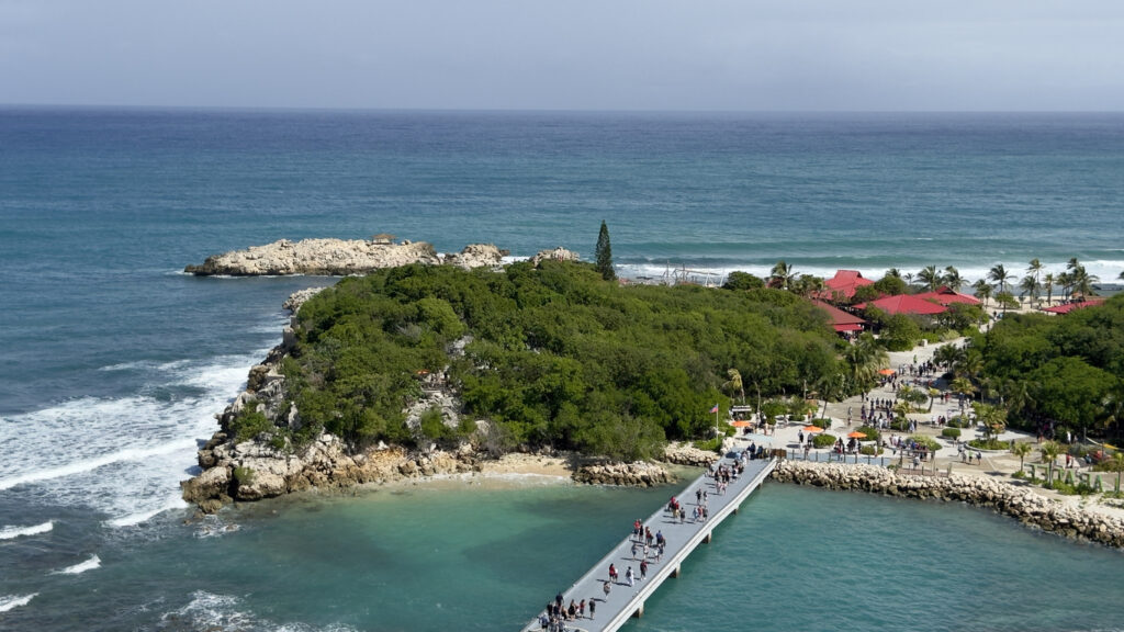 Ein langer Steg führt über türkisfarbenes Wasser zu einer bewaldeten Halbinsel mit roten Dächern.