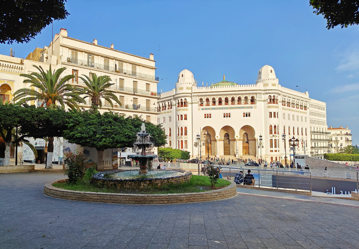 Platzszene in Algier mit Brunnen und weißem Gebäude mit Bögen und kleinen Kuppeln.