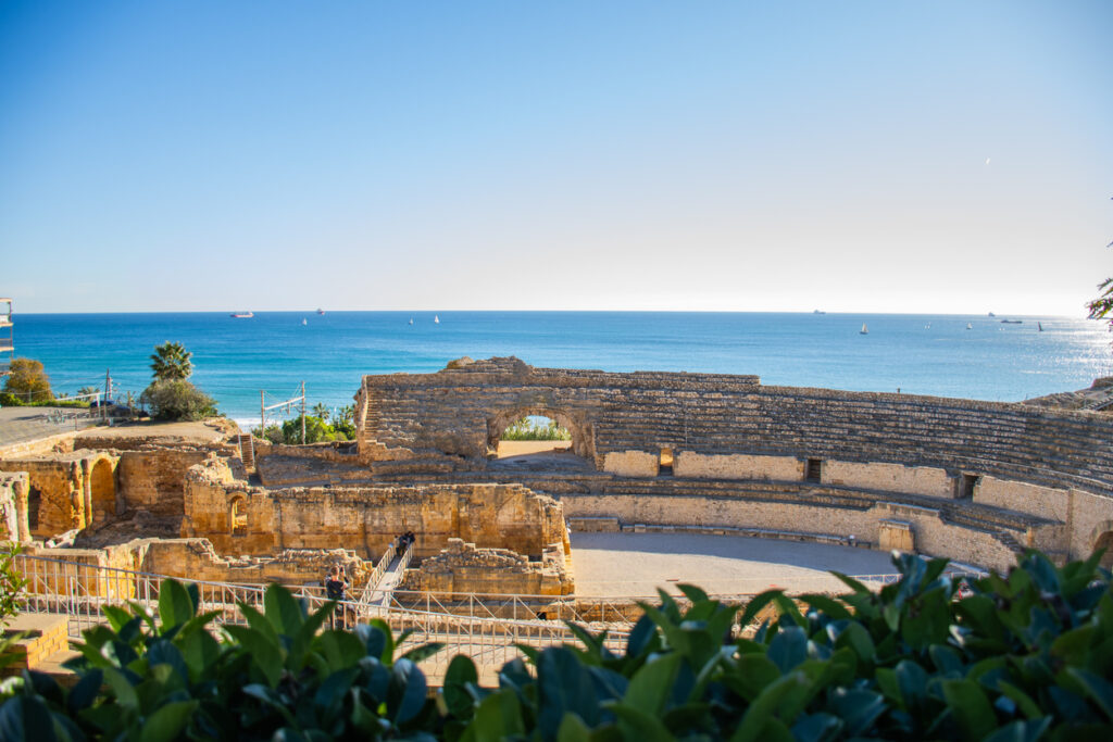 Die steinernen Sitzreihen und die Arena eines antiken Amphitheaters liegen direkt an der Küste unter strahlend blauem Himmel.
