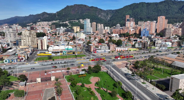 Ein Blick von oben auf eine breite Hauptstraße mit roten Gelenkbussen vor der Skyline von Bogotá und den Anden.