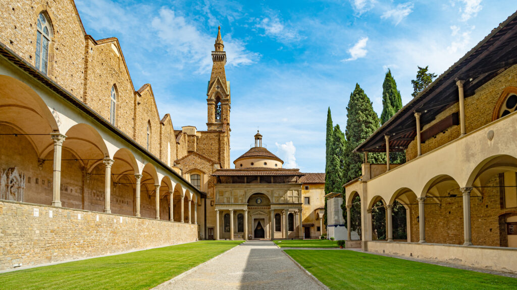 Ein gepflegter grüner Innenhof in Ljubljana mit klassischen Arkadengängen und Blick auf einen spitzen Kirchturm unter blauem Himmel.
