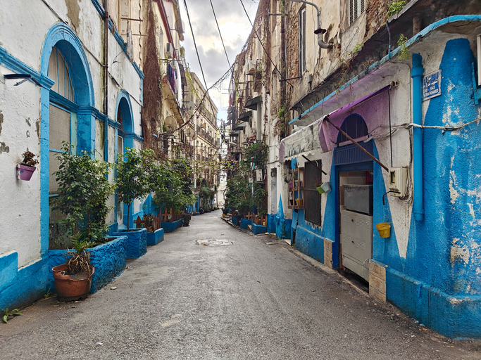 Eine schmale, ansteigende Gasse ist im unteren Bereich der Häuserwände leuchtend blau gestrichen und mit Topfpflanzen dekoriert.