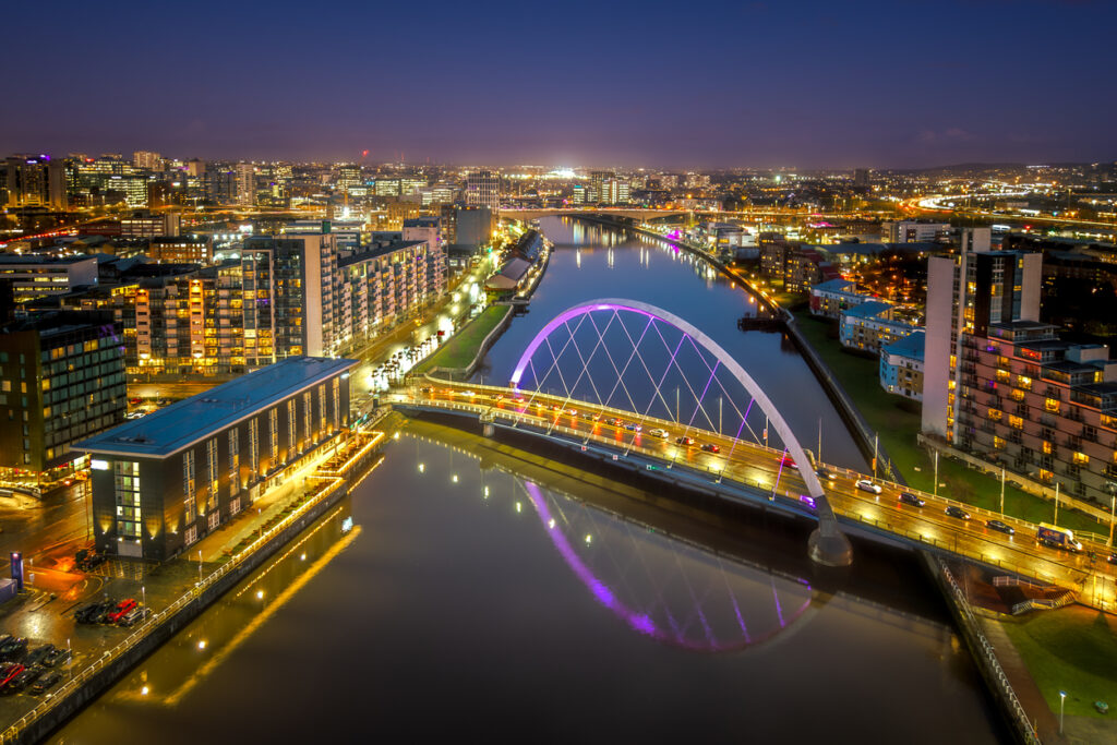 Luftaufnahme einer beleuchteten Bogenbrücke über einen Fluss bei Nacht, deren violetter Lichtbogen sich im Wasser spiegelt, umgeben von modernen Glasgebäuden.