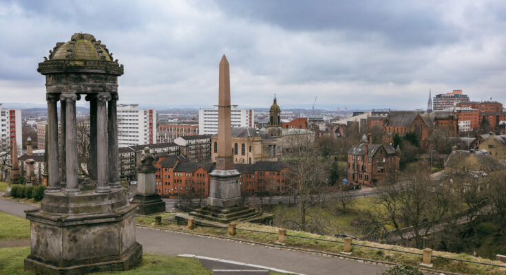 Ein imposantes Denkmal mit einer Säule steht im Vordergrund auf einem grasbewachsenen Hügel, dahinter erstreckt sich das Stadtpanorama von Glasgow unter einem bewölkten Himmel.