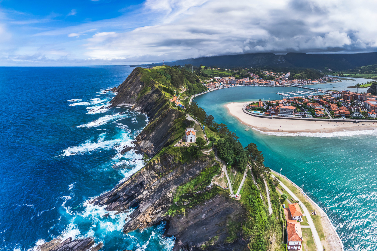 Luftaufnahme einer grünen Landzunge mit steilen Felsen, blauem Ozean, geschützter Bucht und einem Küstenort im Hintergrund.
