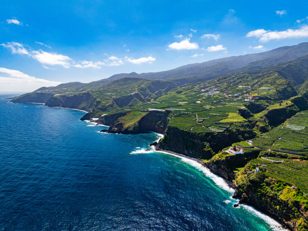 Luftaufnahme der zerklüfteten Nordwestküste von La Palma mit terrassierten grünen Plantagen, steilen Klippen und tiefblauem Meer unter wolkenlosem Himmel.