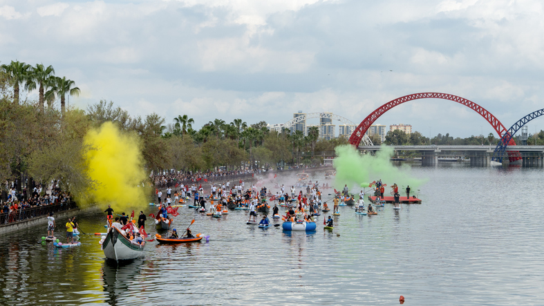 Zahlreiche Menschen auf kleinen Booten und Stand-up-Paddle-Boards feiern auf einem Fluss, während bunter gelber und grüner Rauch aufsteigt.