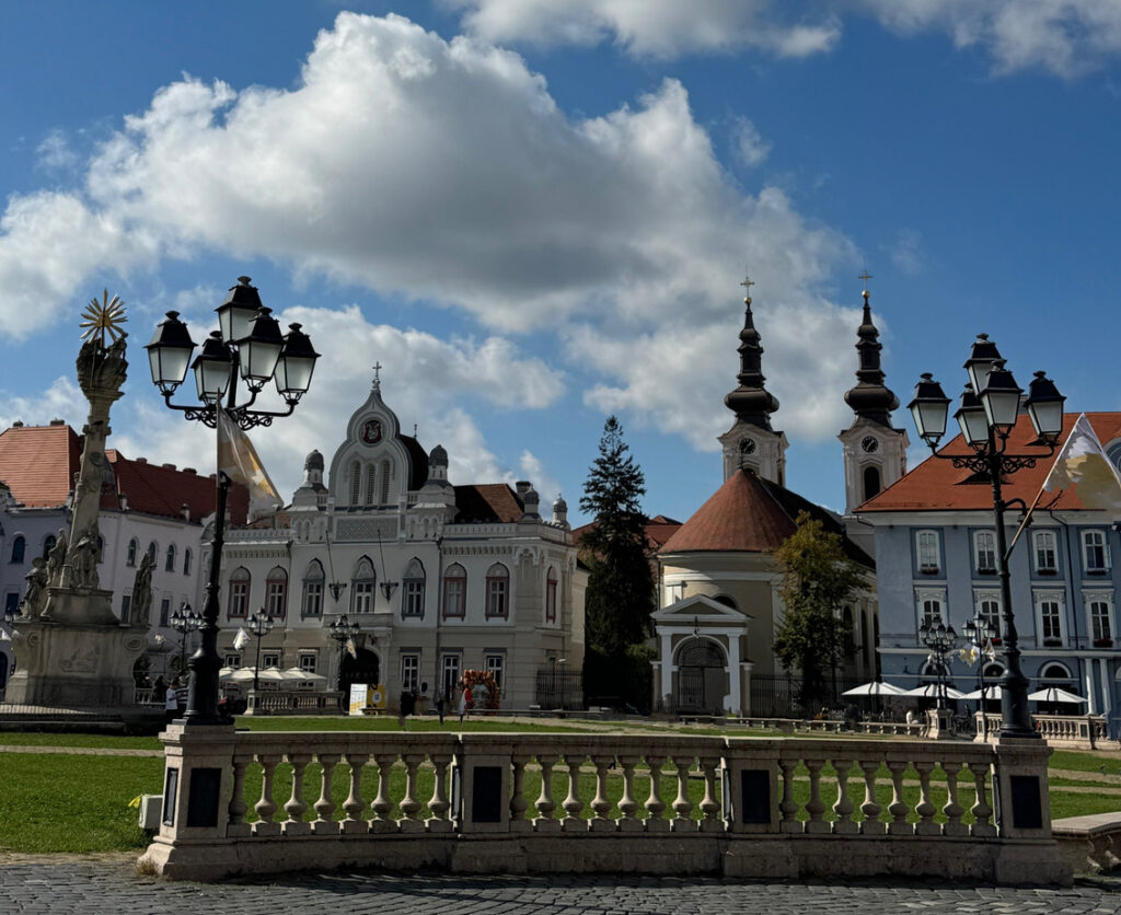 Blick über einen gepflasterten Platz auf prachtvolle barocke Gebäude und eine Kirche mit zwei dunklen Zwiebeltürmen unter bewölktem Himmel.
