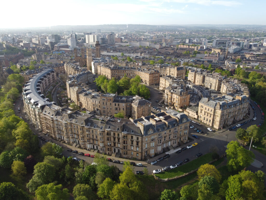 Luftbild einer halbkreisförmig angeordneten Häuserzeile aus hellem Stein mit dunklen Dächern, umgeben von dichten grünen Bäumen und dem Stadtpanorama.