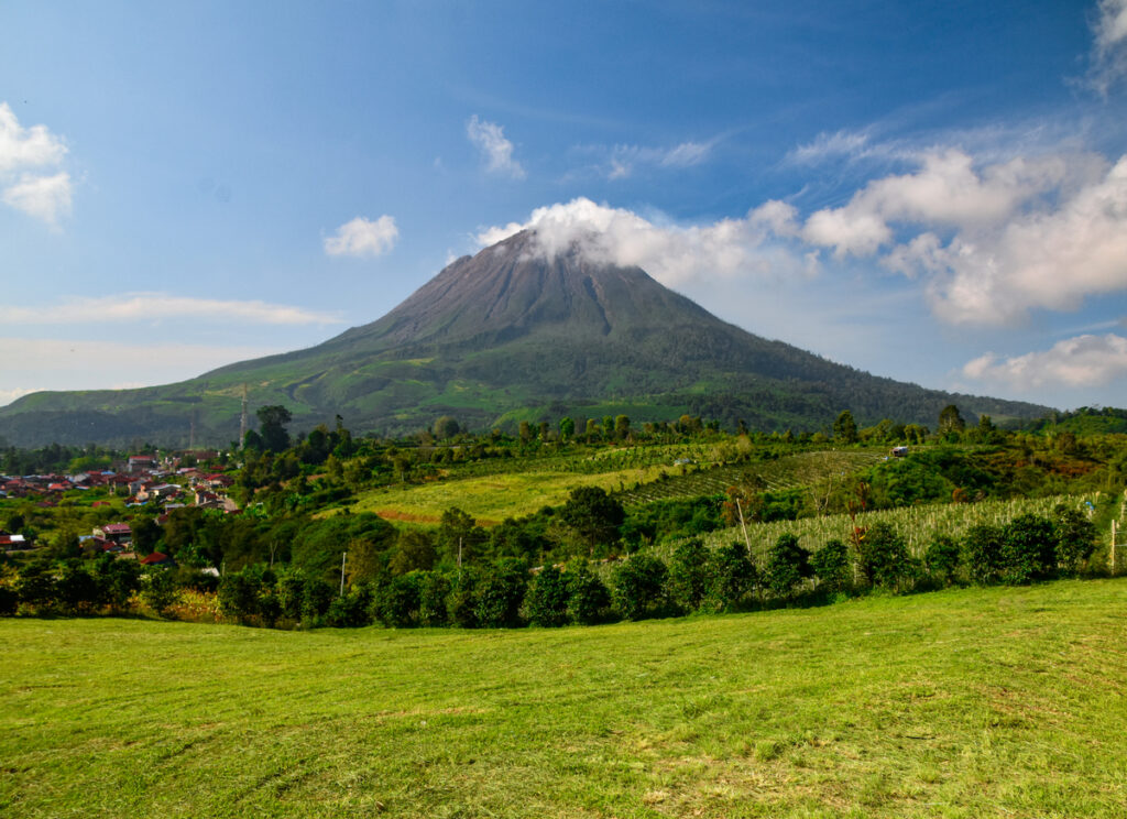 Ein großer Vulkan mit einer kleinen Wolke am Gipfel thront über grünen Feldern und einer kleinen Stadt.