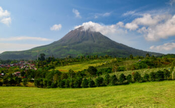 Ein großer Vulkan mit einer kleinen Wolke am Gipfel thront über grünen Feldern und einer kleinen Stadt.