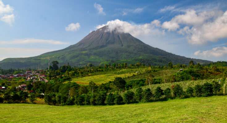 Ein großer Vulkan mit einer kleinen Wolke am Gipfel thront über grünen Feldern und einer kleinen Stadt.