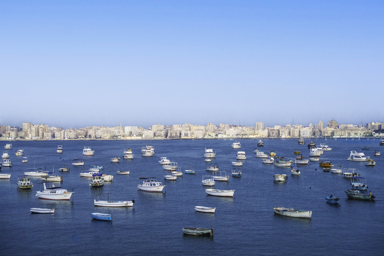 Zahlreiche kleine weiße Fischerboote und Yachten ankern auf dem tiefblauen Wasser vor der hellen Kulisse der dicht bebauten Stadtfront unter strahlend blauem Himmel.