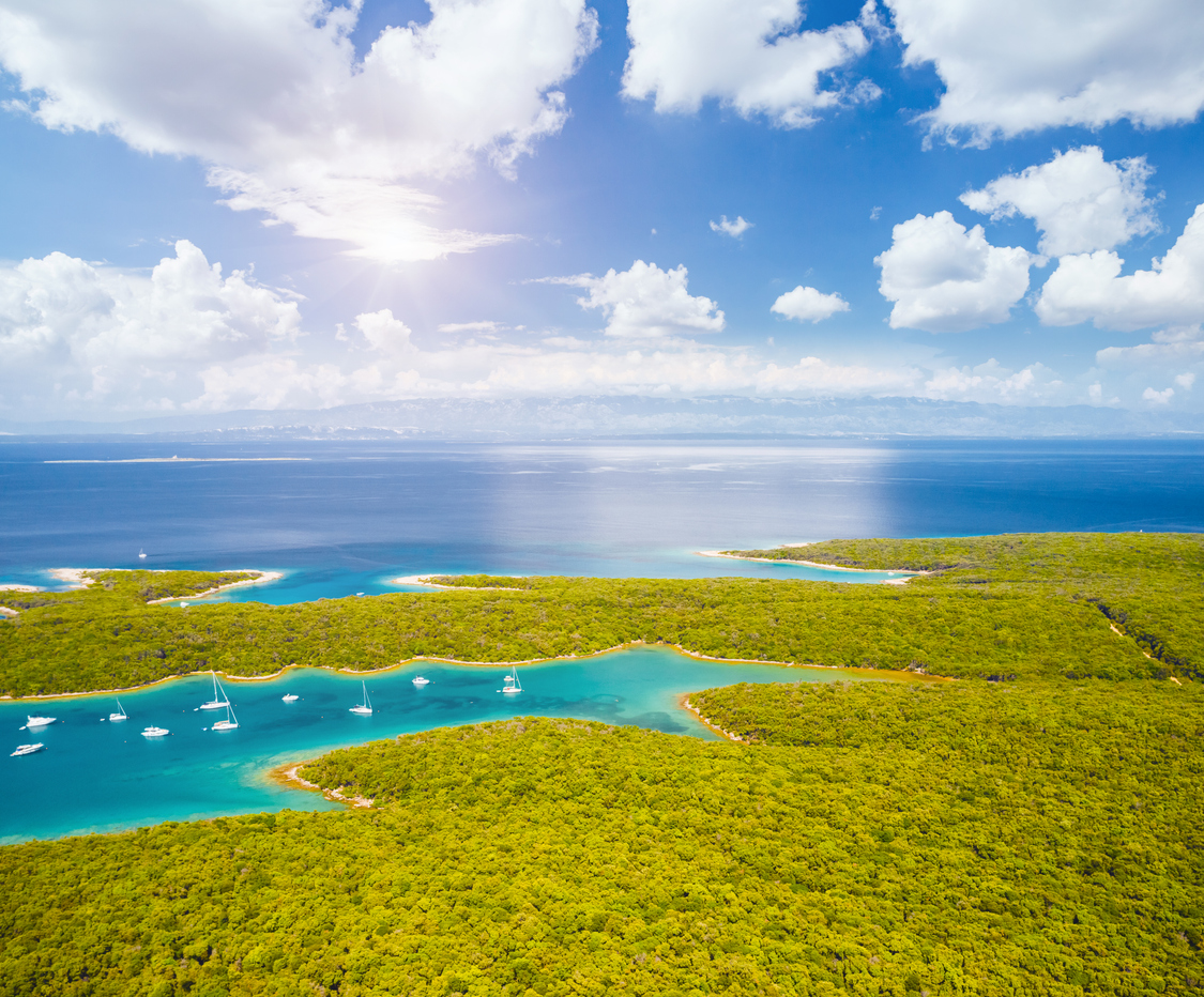 Eine Drohnenaufnahme zeigt eine tiefblaue Meeresbucht mit ankernden Jachten, umgeben von einem dichten, grünen Teppich aus mediterranen Wäldern.