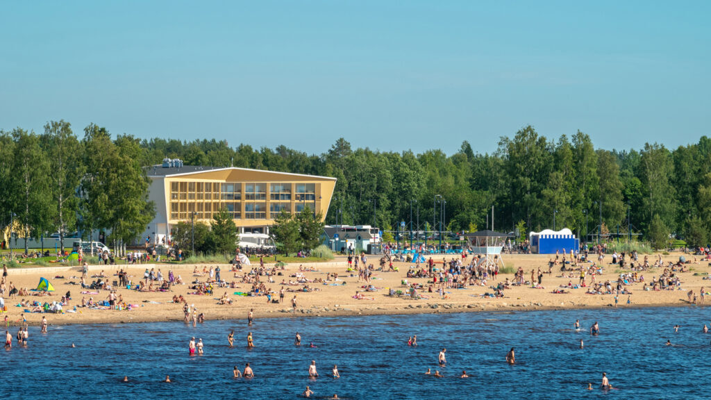 Viele Menschen entspannen am Sandstrand und baden im Wasser, während dahinter ein helles Gebäude und Bäume stehen.