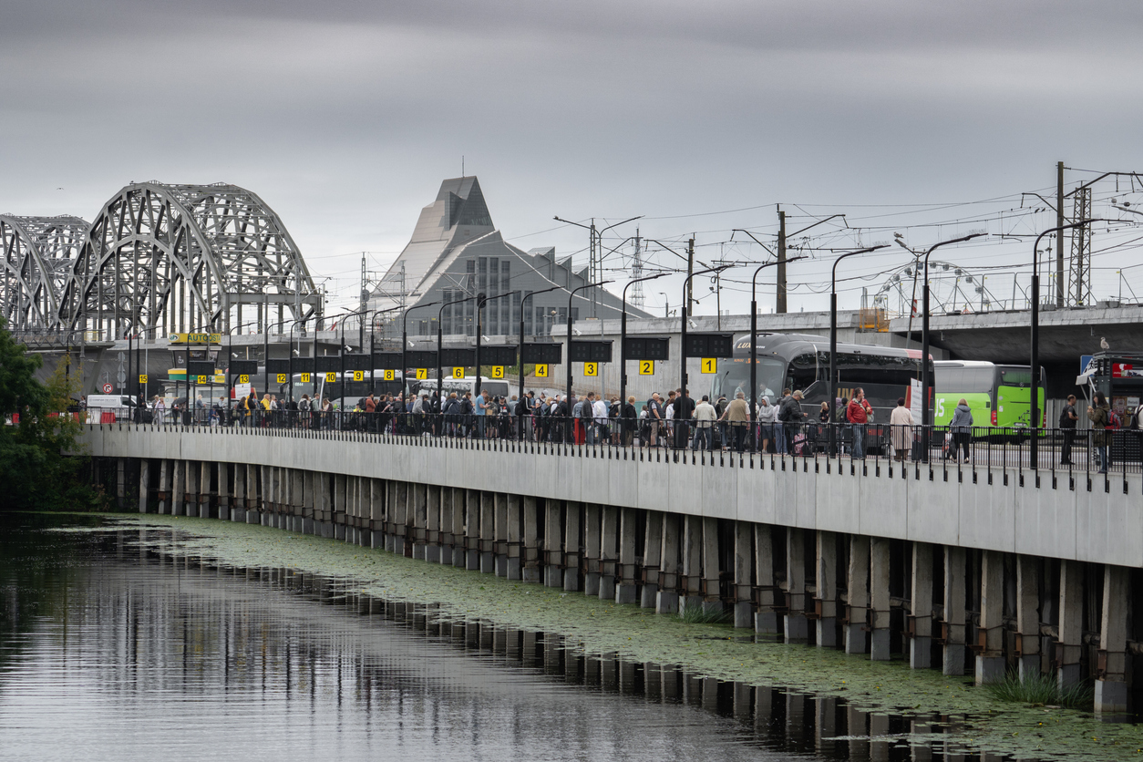Menschen warten an einem Busbahnhof am Flussufer vor der Kulisse der Nationalbibliothek und der Eisenbahnbrücke.