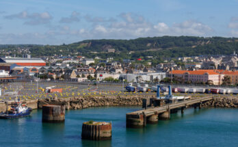 Ein Panorama des Hafengeländes mit LKWs, einem Schlepper und der Stadt im Hintergrund vor grünen Hügeln.