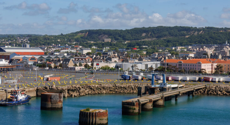 Ein Panorama des Hafengeländes mit LKWs, einem Schlepper und der Stadt im Hintergrund vor grünen Hügeln.