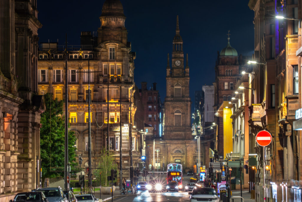Eine nächtliche Straßenszene in Glasgow mit beleuchteten historischen Sandsteingebäuden, einem Doppeldeckerbus und hellen Autoscheinwerfern im Vordergrund.