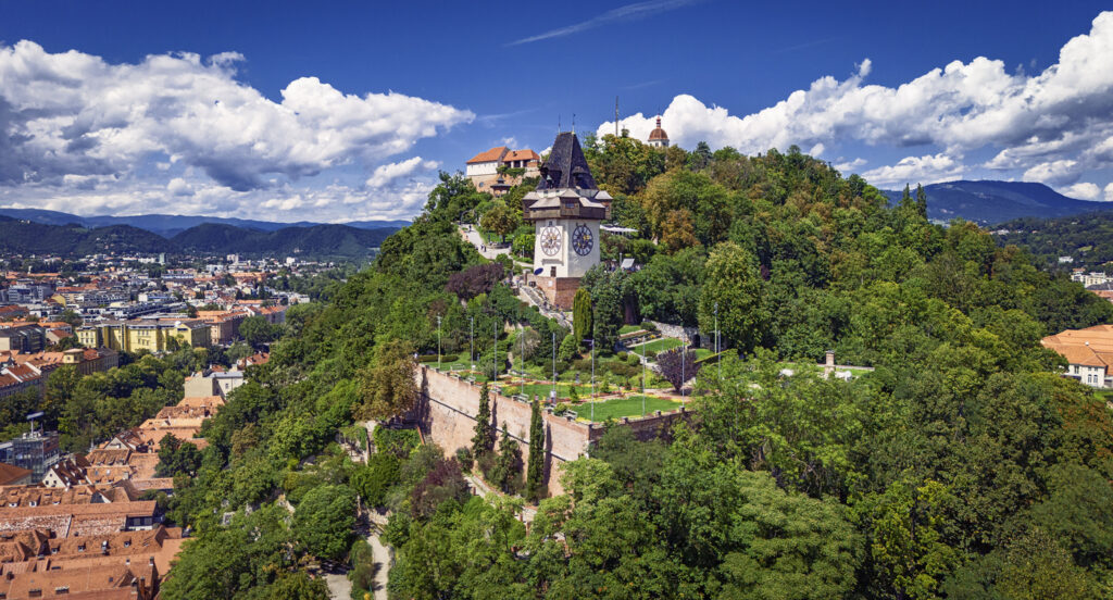 Eine Luftaufnahme vom Schlossberg in Graz, auf dem der Uhrturm inmitten von dichten Bäumen und historischen Mauern thront.