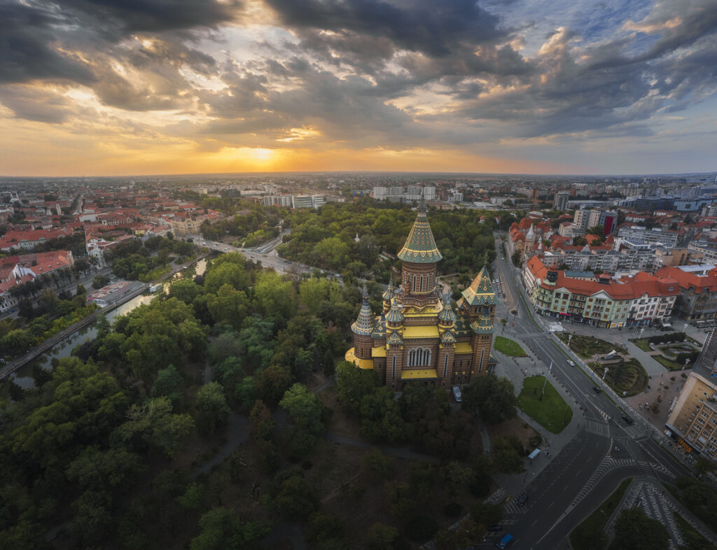 Ein Luftbild zeigt die Kathedrale inmitten dichter grüner Bäume, umgeben von den roten Dächern der umliegenden Stadtviertel während des Sonnenuntergangs.